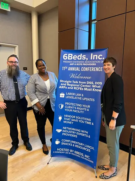 Two diverse women and a man stand on each side of a large floor sign describing the event called 6beds Annual conference