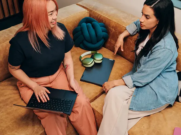 women holding laptops face each other on a corner sofa talking