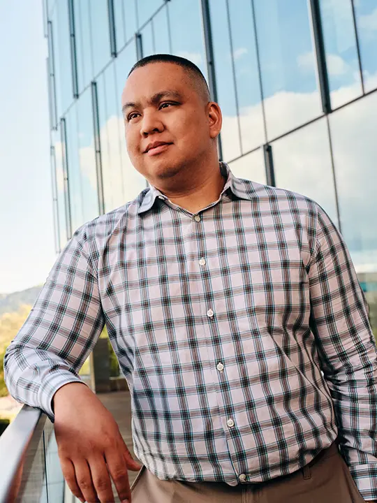 man wearing plaid dress shirt stands on a terrace outdoors with a glass mirrored building in background