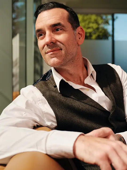 man in dress shirt and vest sits on sofa smiling looking out sunny window